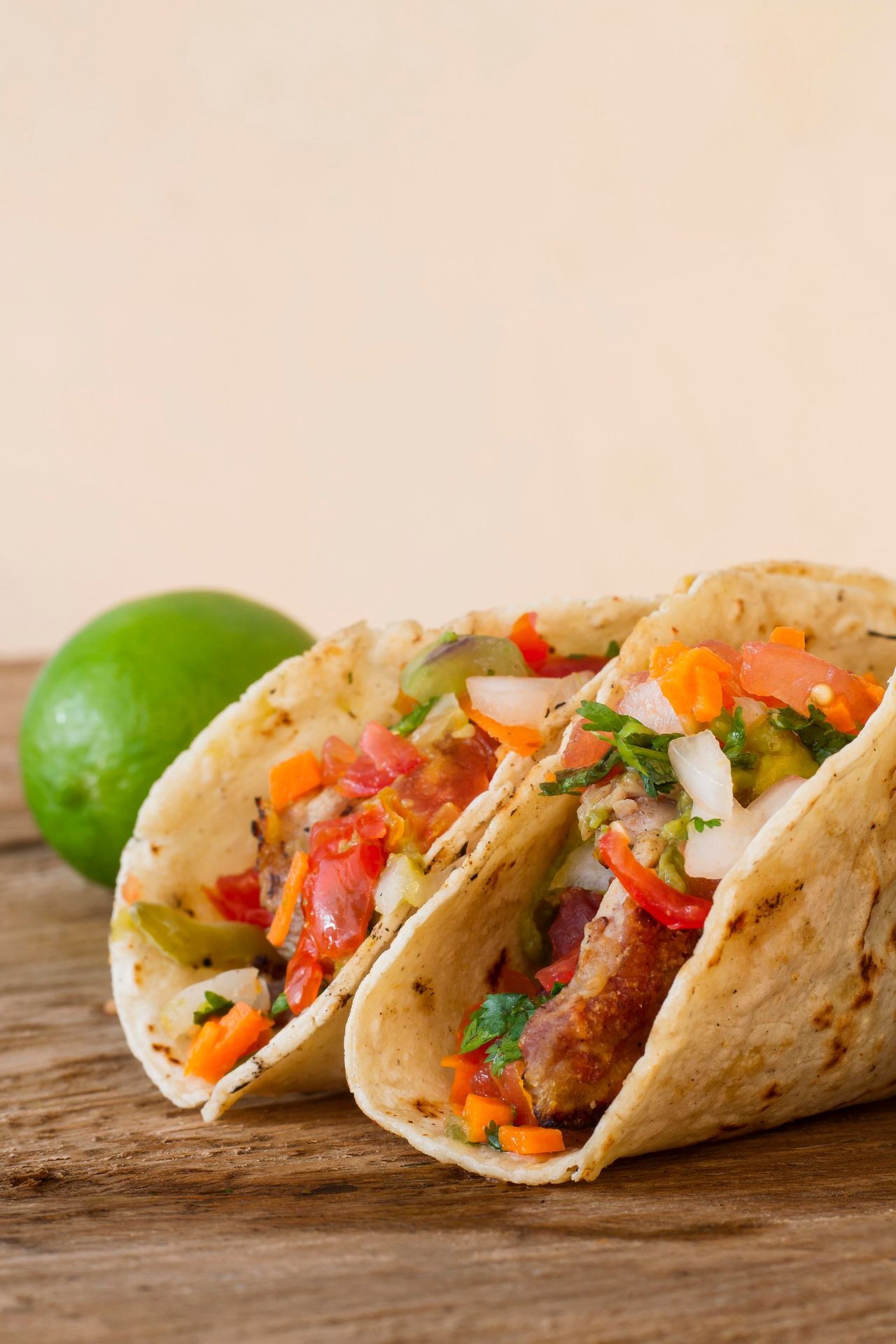 Two tacos filled with colorful vegetables and meat served with a lime on a wooden surface.